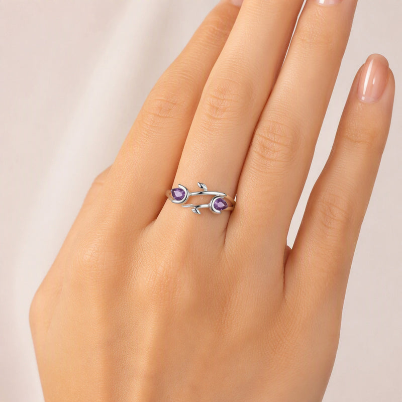 A silver ring with two small purple gemstones and leaf-like details, shown worn on a person's finger against a soft light background.