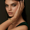 A close-up portrait of a woman with dark hair and green eyes wearing a chunky gold chain-link bracelet on her wrist, posed against a dark background.