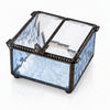 A square glass jewelry box with a hinged lid, featuring light blue textured glass sides, clear glass panels on top, and a dark metal frame with a butterfly ornament on the lid.