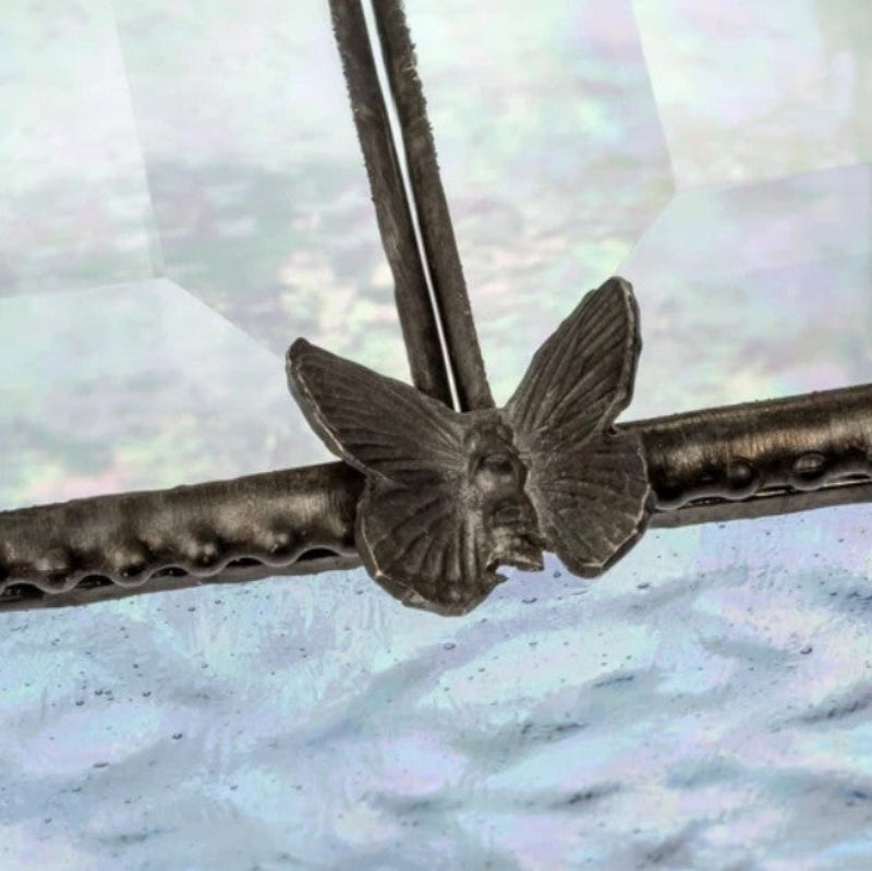 A close-up of a dark metallic butterfly-shaped decorative element attached to a textured horizontal bar, with a soft iridescent blue-white background.