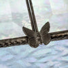 A close-up of a dark metallic butterfly-shaped decorative element attached to a textured horizontal bar, with a soft iridescent blue-white background.