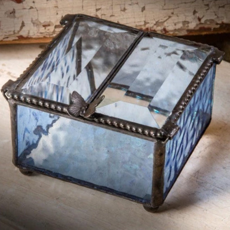 A small decorative glass jewelry box with a hinged lid, featuring blue-tinted glass panels, a dark metal frame with bead detailing, and a butterfly ornament on the lid, set on a rustic wooden surface.