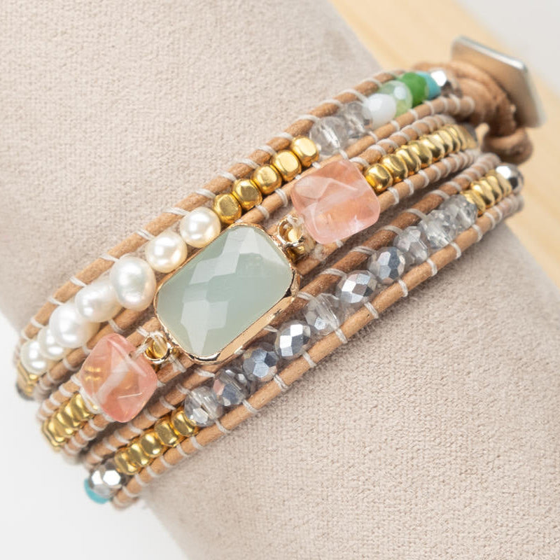 A close-up of a multi-strand beaded wrap bracelet displayed on a beige fabric jewelry stand. The bracelet features tan cords, gold and silver metallic beads, white faux pearls, faceted clear rhinestones, peach-colored stone pieces, and a central light green rectangular stone.